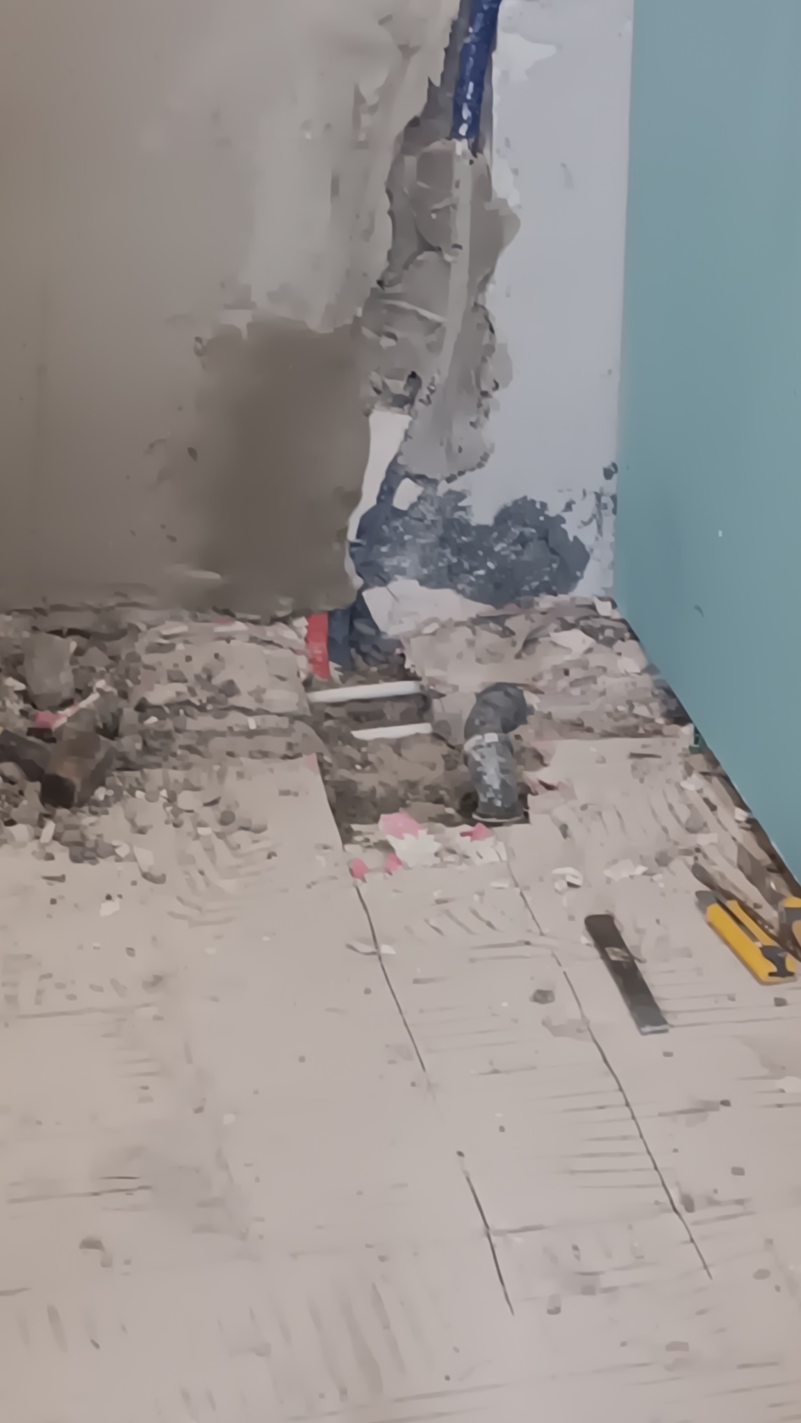 Fragment zdemontowanej łazienki z odsłoniętymi rurami, gruzem i narzędziami na podłodze, widoczne ślady po usuniętej glazurze na ścianie.