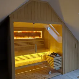 Sauna wkomponowana w poddasze ze skośnym sufitem, oświetlona od wewnątrz, z przeszklonymi drzwiami, widoczne kamienie solne na ścianie i akcesoria saunowe.