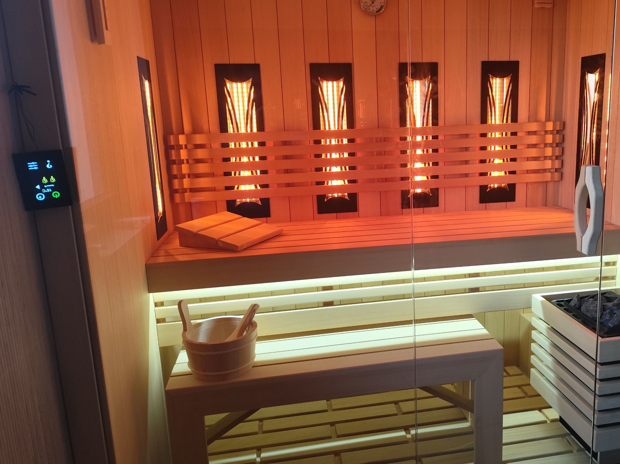 Wnętrze sauny z drewnianymi ławkami, oświetleniem podczerwienią i panelem sterowania po lewej stronie, widoczny drewniany ceber z chochlą i pojemnik na kamienie