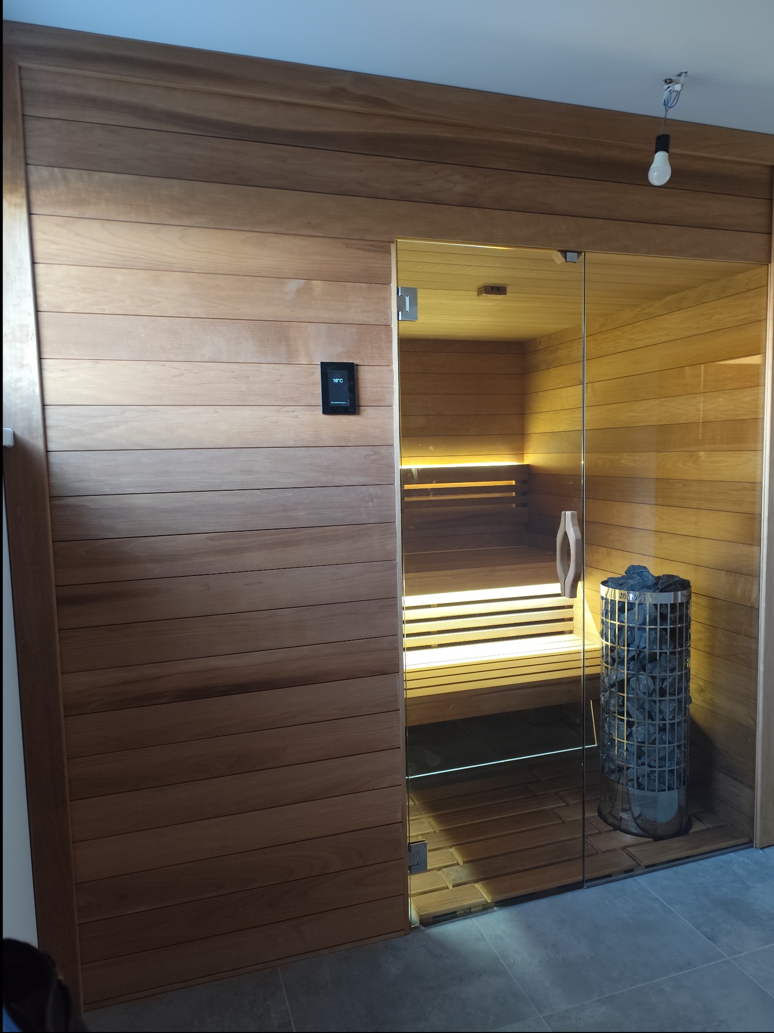 Nowoczesna sauna z drewnianym wykończeniem, szklanymi drzwiami, oświetleniem LED pod ławkami i piecem z kamieniami w metalowej obudowie. Na ścianie zewnętrznej widoczny panel sterowania temperaturą.