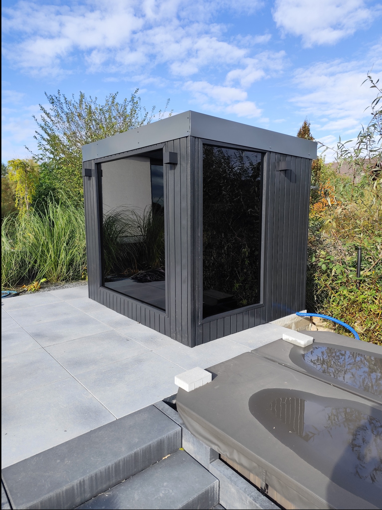 Nowoczesna, ciemnoszara kabina z dużymi oknami, prawdopodobnie sauna, ustawiona na szarym tarasie z płyt betonowych, obok przykryte jacuzzi, otoczona zielenią.