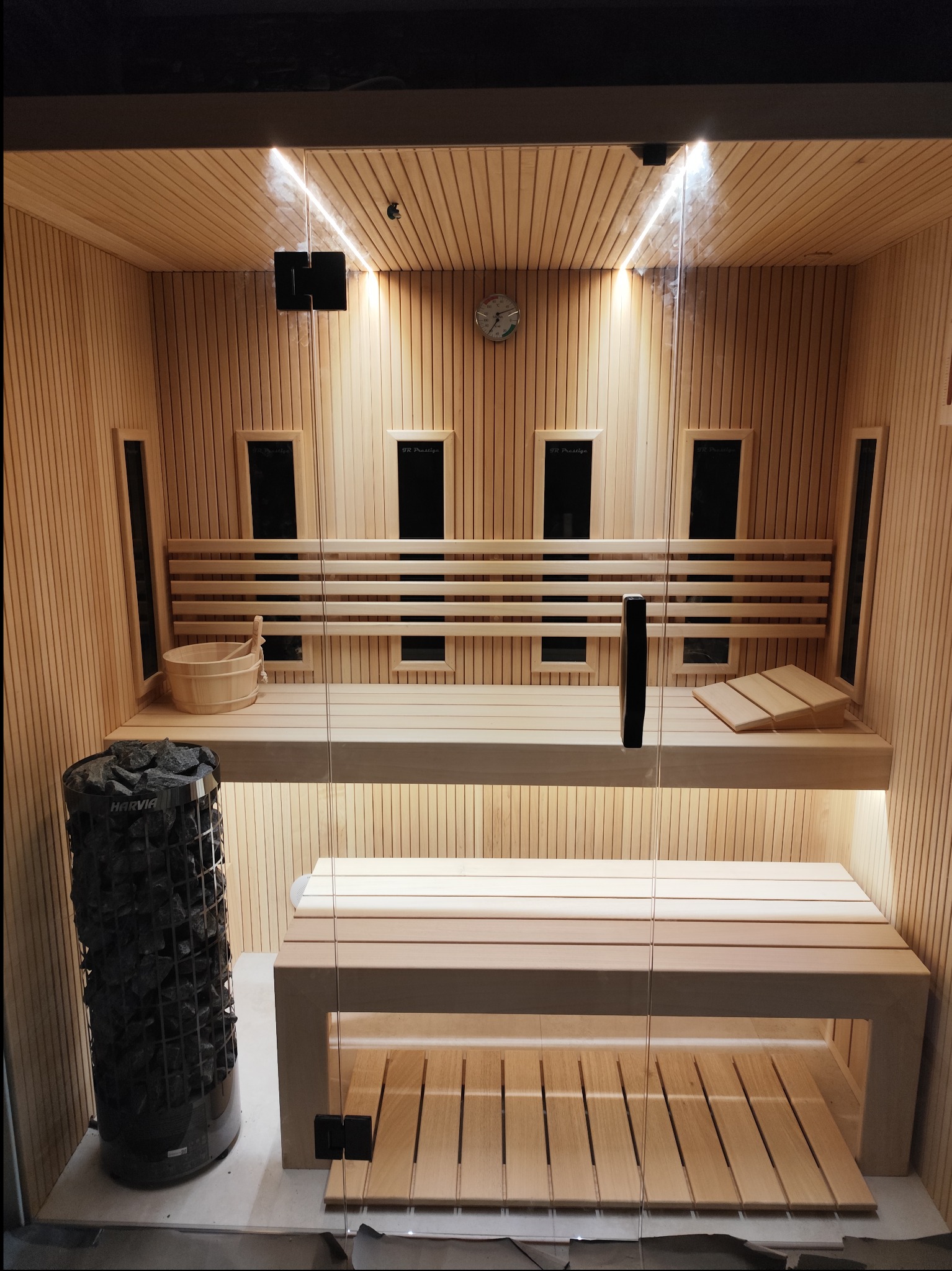 Wnętrze nowej sauny z drewnianymi panelami ściennymi, dwupoziomowymi ławkami, piecem z kamieniami i szklanymi drzwiami.