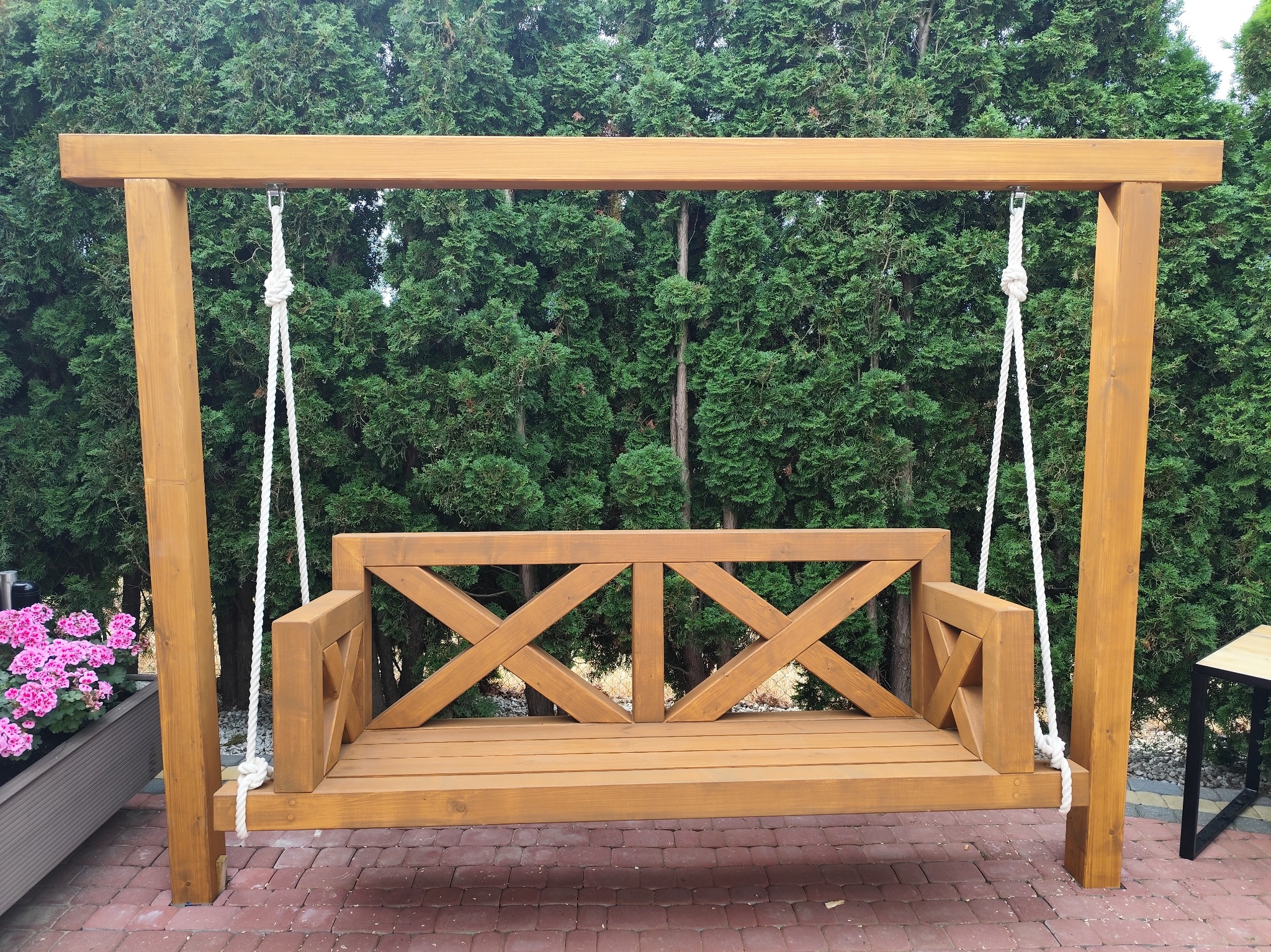 Drewniana huśtawka ogrodowa zawieszona na linach, z oparciem z motywem krzyża, na tle zielonych tuj i czerwonej kostki brukowej, z donicą z różowymi kwiatami po lewej stronie.