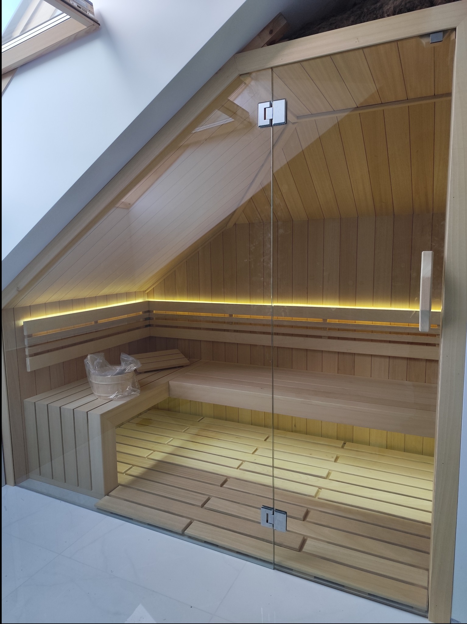Nowoczesna sauna w poddaszu z przeszklonymi drzwiami, drewnianymi ławkami i podświetleniem LED, wiadro na wodę w rogu.