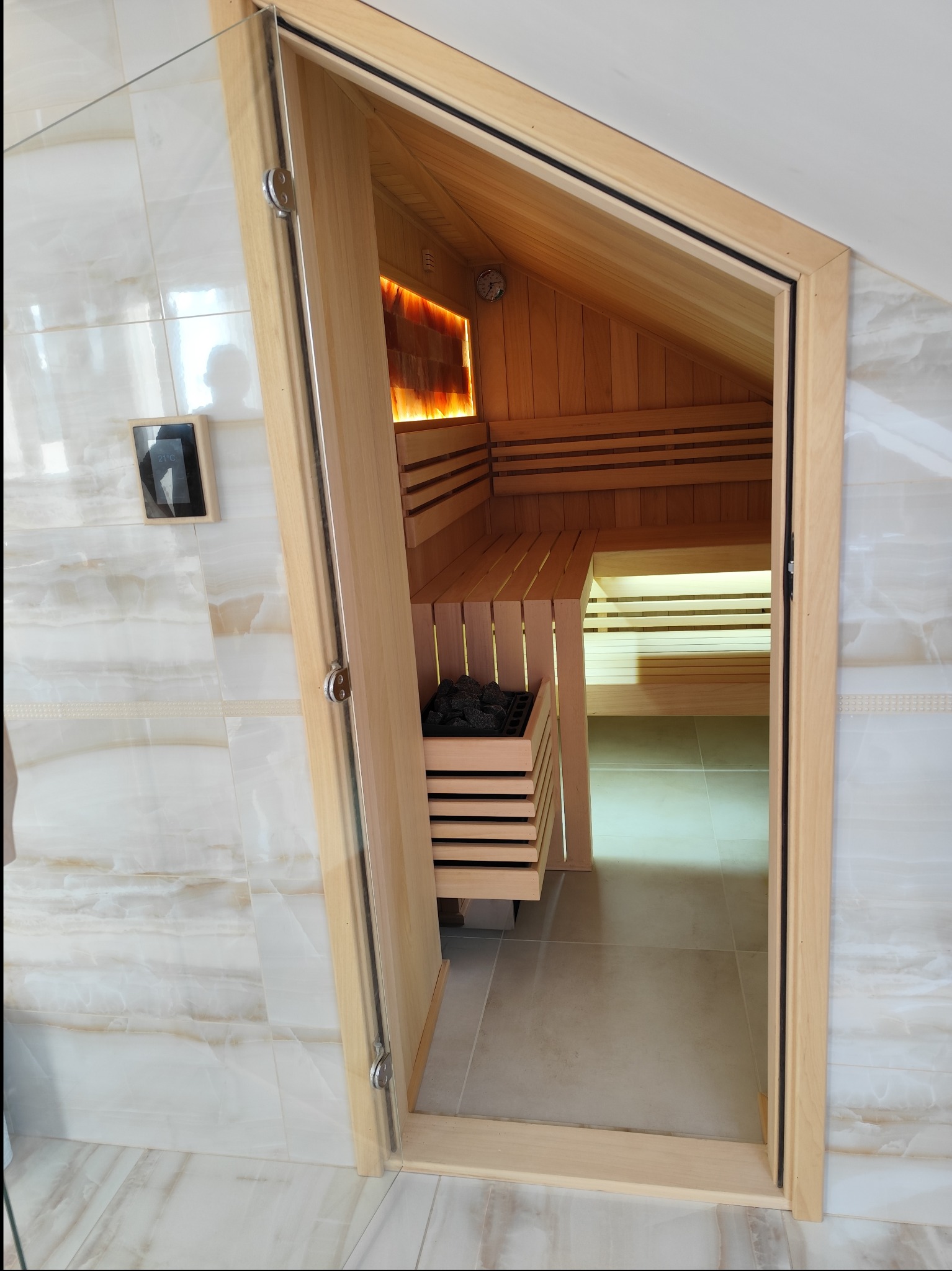 Wnętrze nowej sauny z drewnianymi ławkami, piecem na kamienie i oświetleniem solnym, widoczne przez otwarte szklane drzwi na tle marmurowych płytek.