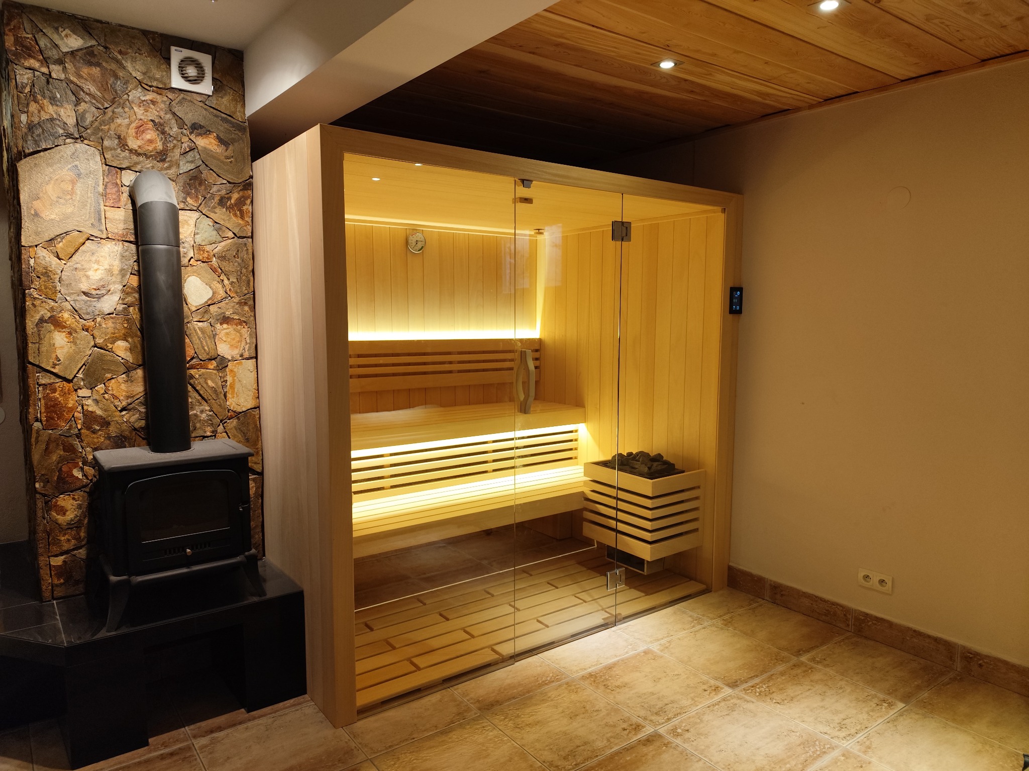 Nowoczesna sauna z przeszklonymi drzwiami, oświetleniem LED i piecem opalanym drewnem, umieszczonym przy ścianie wyłożonej kamieniem.