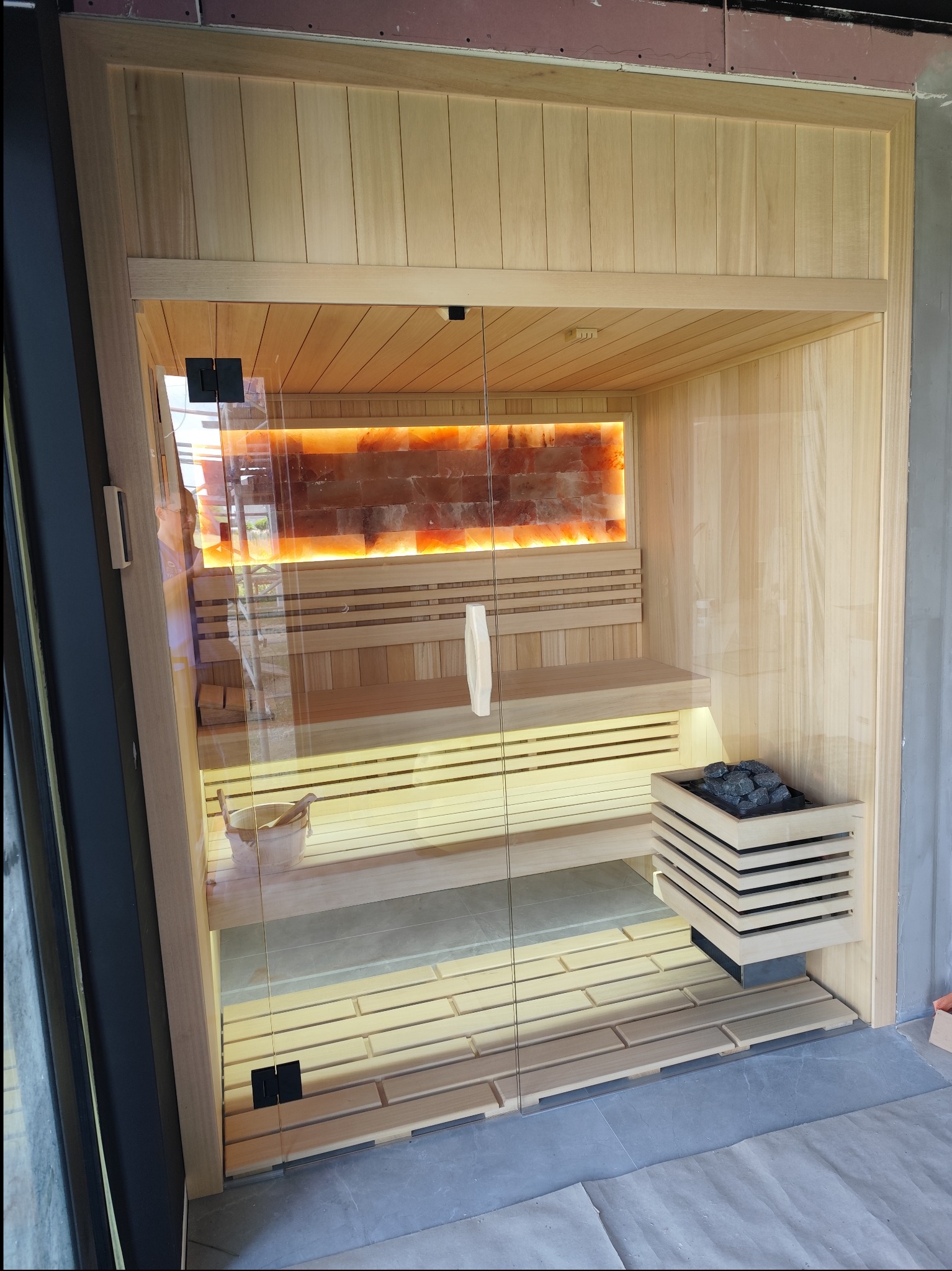 Wnętrze sauny z drewnianym wykończeniem, oświetloną ścianą z soli i szklanymi drzwiami, widoczne kamienie do sauny w drewnianej obudowie oraz drewniane wiadro z chochelką.