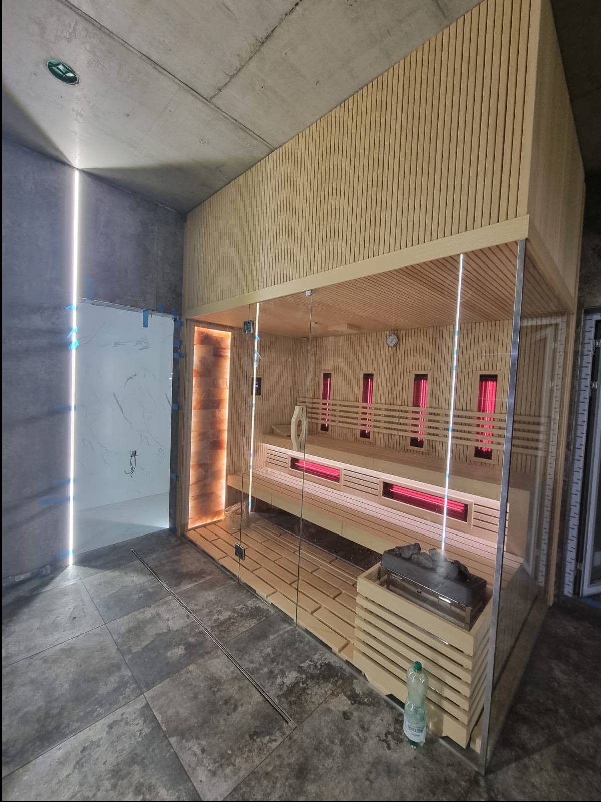 Nowoczesna sauna z przeszklonymi ścianami, drewnianym wykończeniem i elementami podświetlenia, widoczne ławy, piec z kamieniami i butelka wody na podłodze.