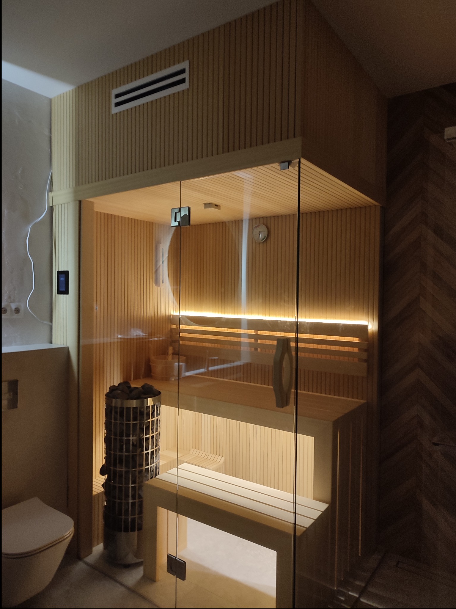 Wnętrze sauny z piecem na kamienie, drewnianymi ławkami i przeszklonymi drzwiami, widoczny panel sterowania na ścianie i fragment toalety obok.