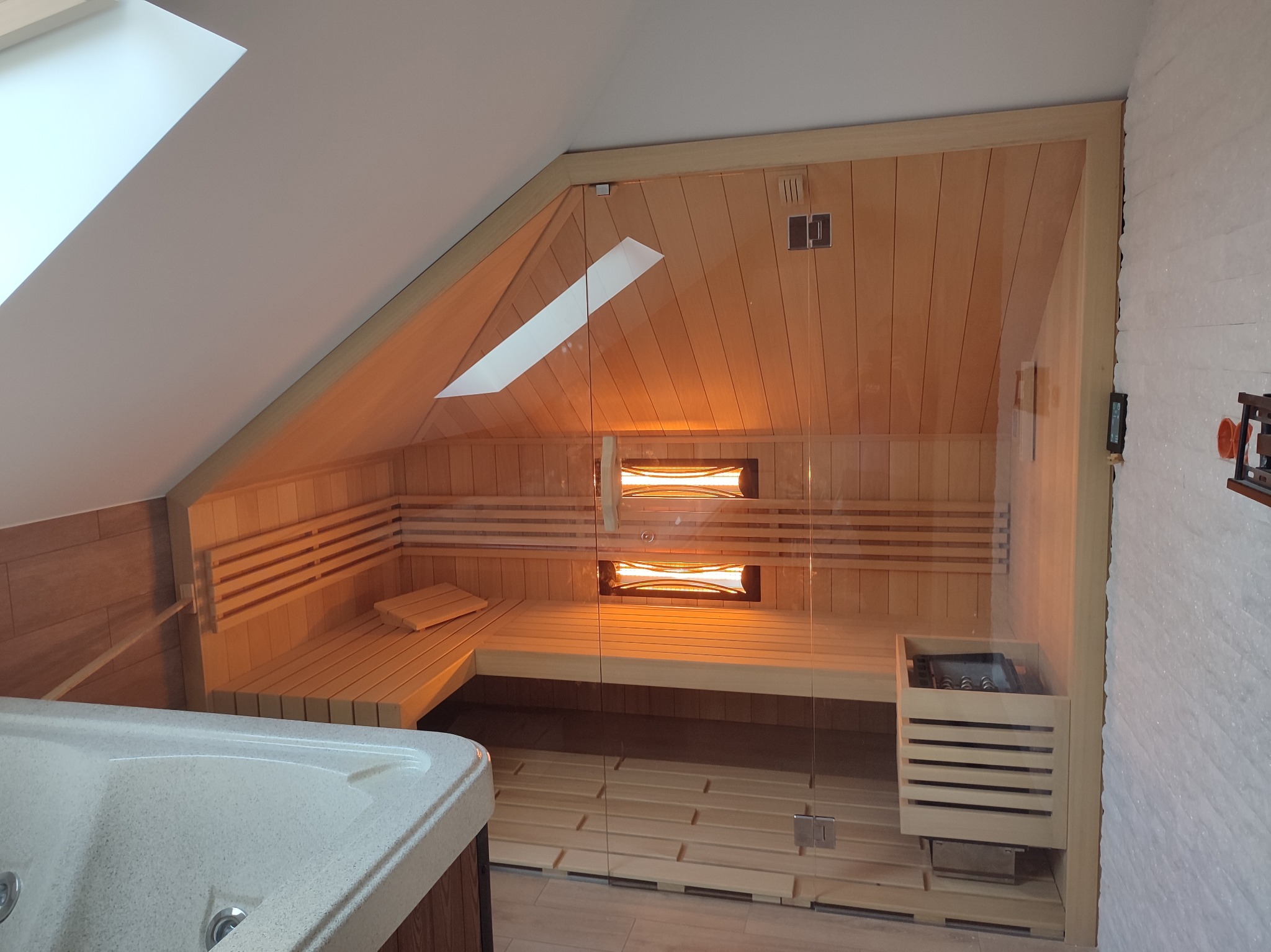 Sauna w pomieszczeniu ze skośnym sufitem, widoczna przez szklane drzwi, obok wanna z hydromasażem, drewniane wykończenie i oświetlenie wewnętrzne.