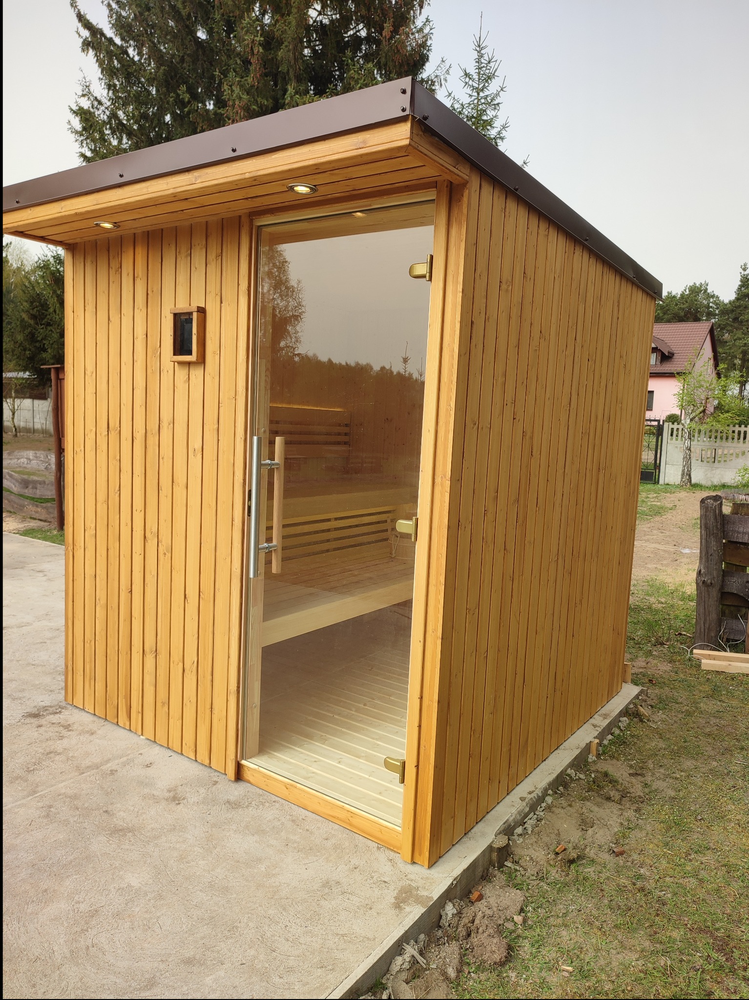 Gotowa, zewnętrzna, drewniana konstrukcja z przeszklonymi drzwiami, prawdopodobnie sauna, z oświetleniem punktowym w dachu, na betonowej wylewce, z widocznym fragmentem trawnika i drzew w tle.