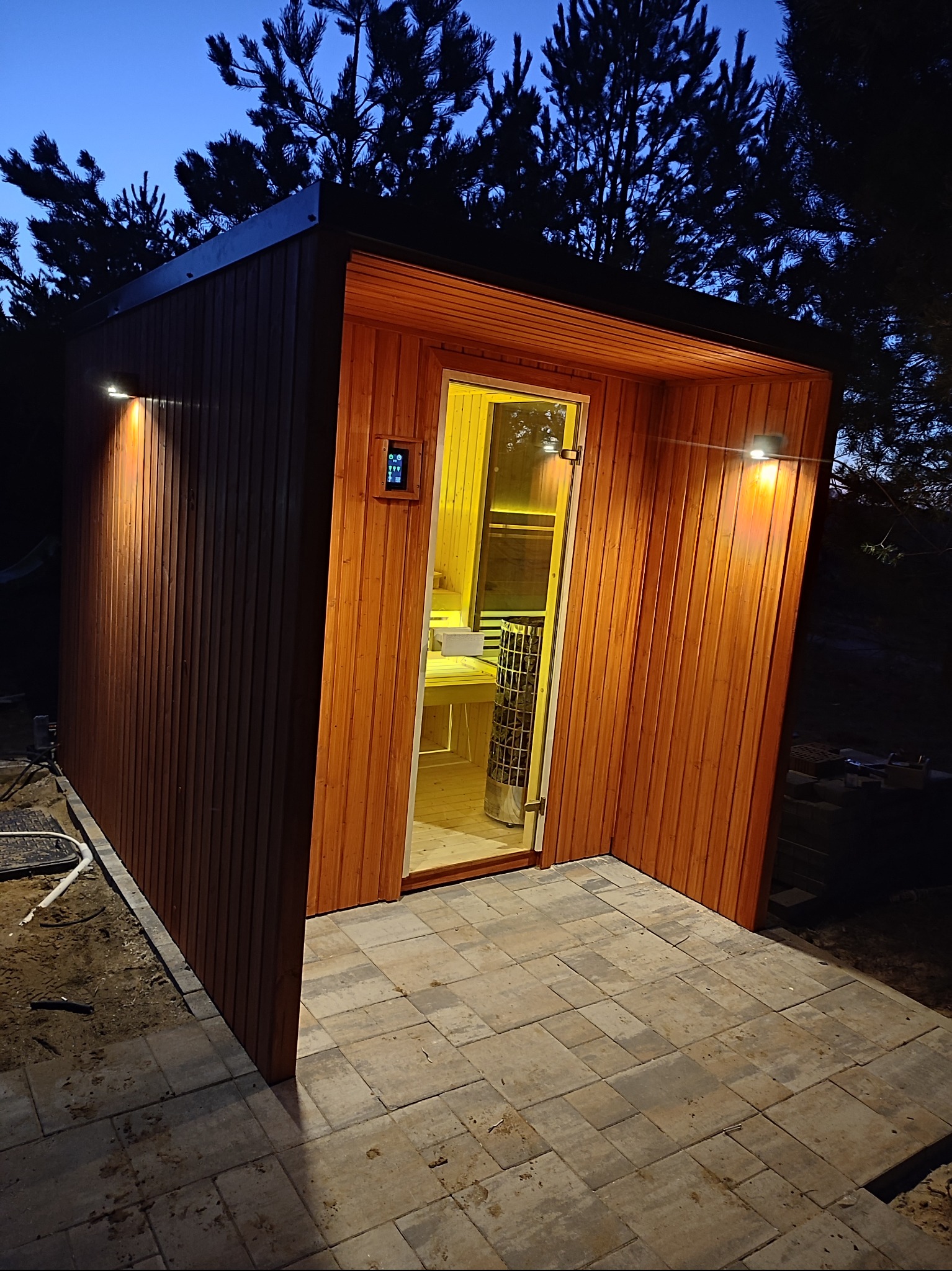 Nowoczesna, drewniana sauna zewnętrzna z podświetlaną elewacją i przeszklonymi drzwiami, widoczny piec wewnątrz, otoczona kostką brukową i drzewami w wieczornym oświetleniu.