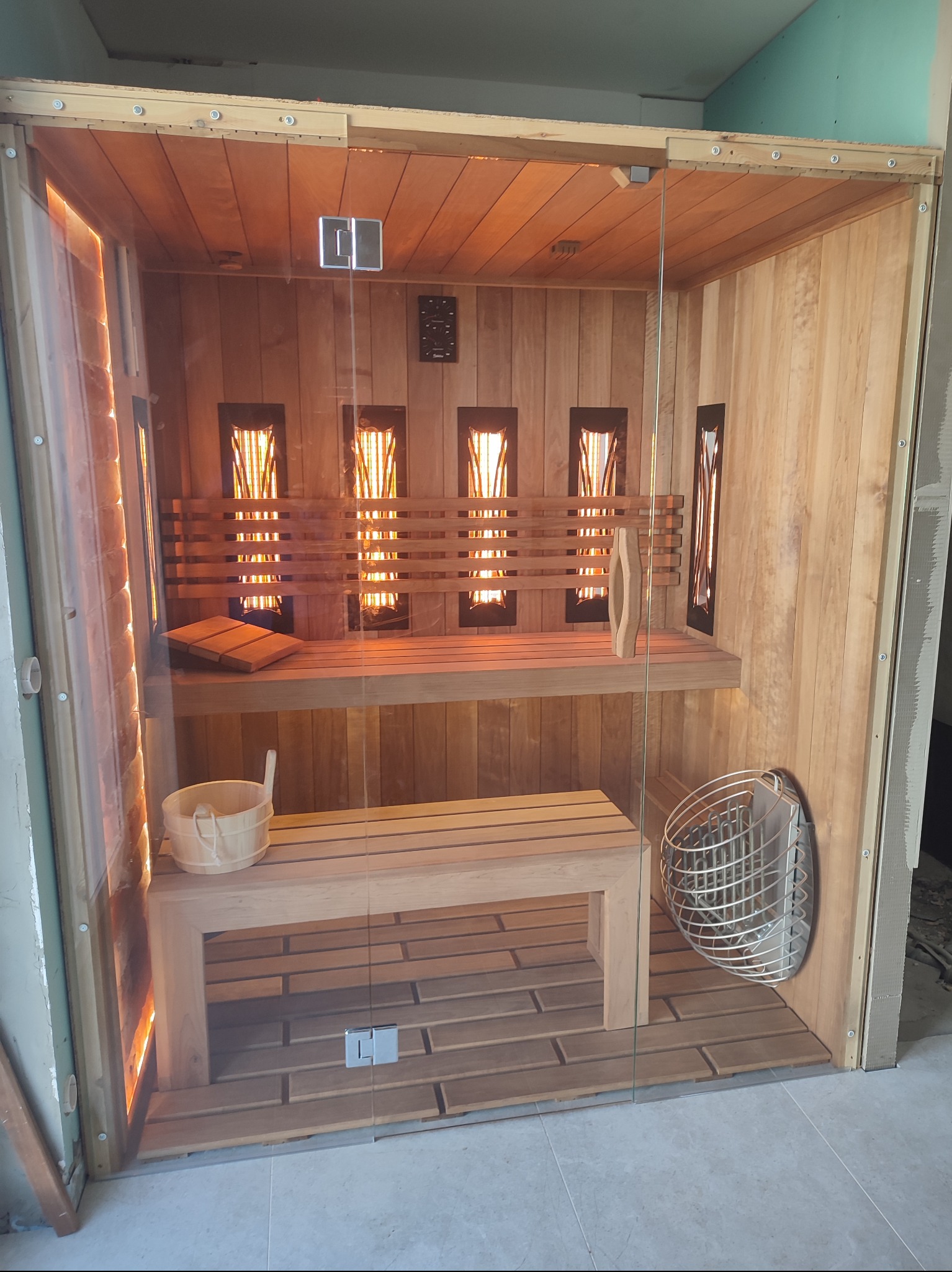 Nowoczesna sauna z drewnianym wykończeniem i szklanymi drzwiami, widoczne drewniane ławki, oświetlenie halogenowe i metalowy piec, w tle surowy stan wykończenia pomieszczenia.