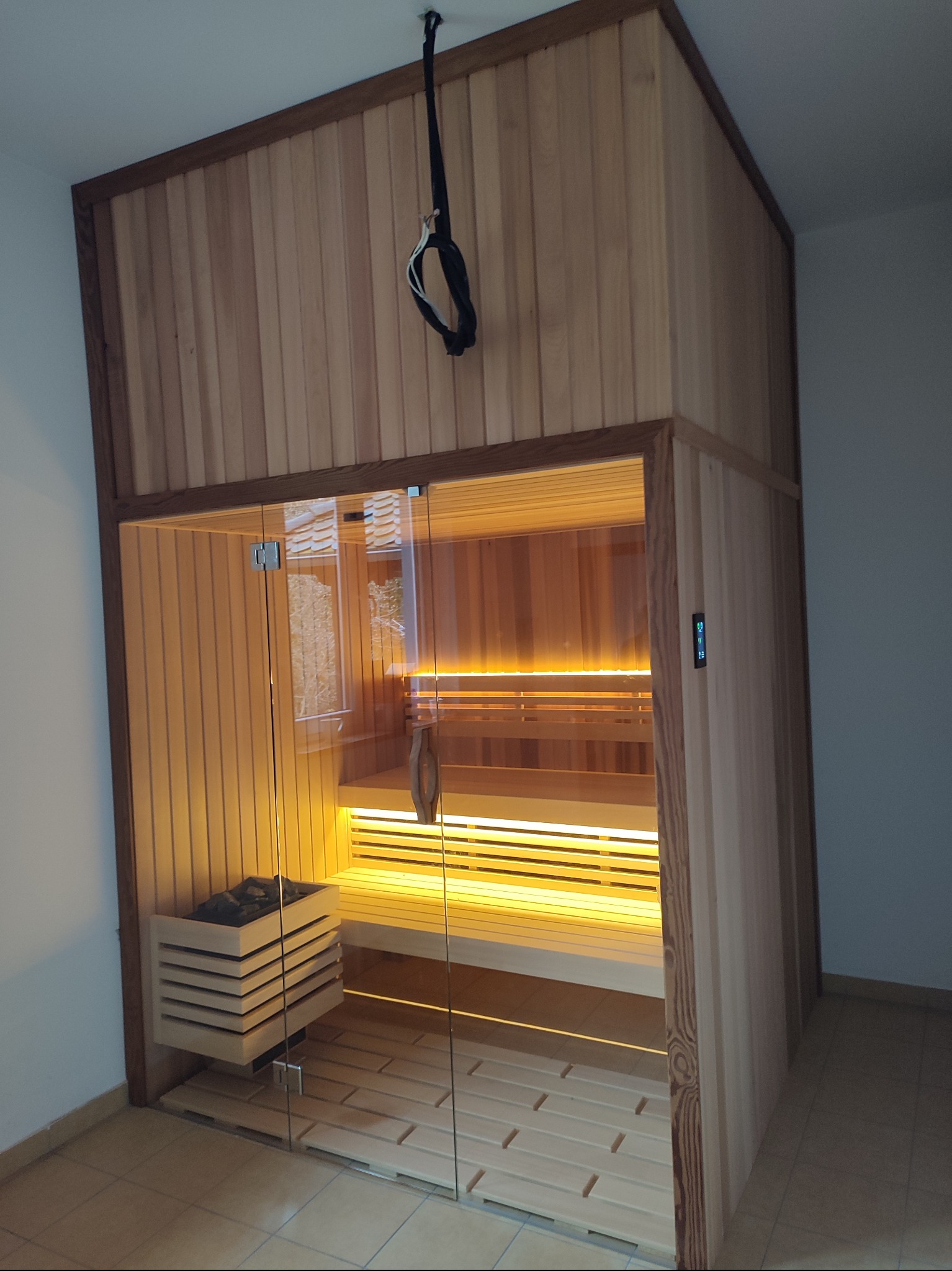 Wnętrze nowoczesnej sauny z jasnego drewna, z przeszklonymi drzwiami, oświetleniem LED i piecem na kamienie, widoczne elementy sterowania na ścianie.