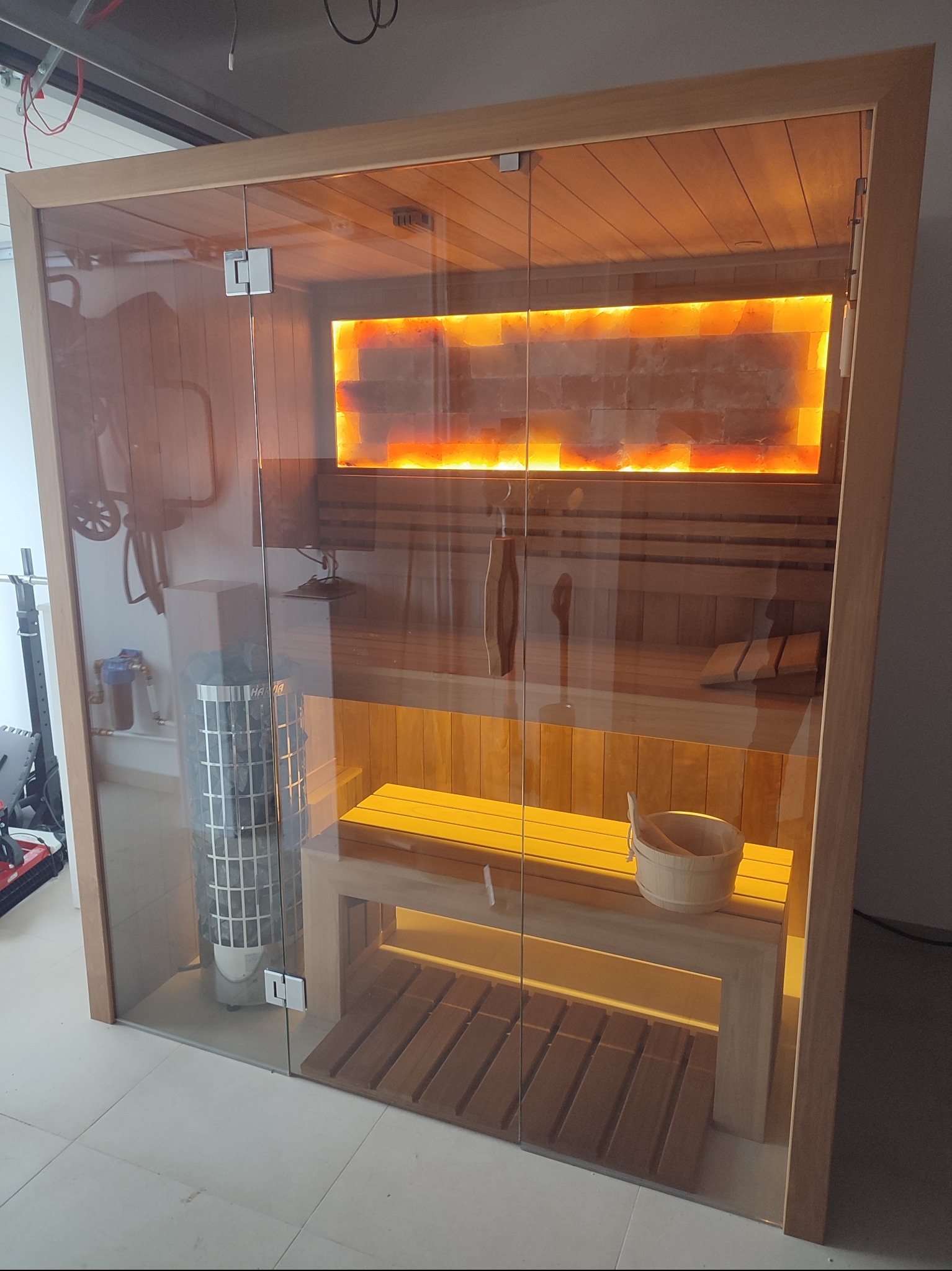 Szklana sauna z drewnianym wykończeniem, podświetlona ściana z cegieł solnych, piec na kamienie i drewniane ławy, widok z zewnątrz.