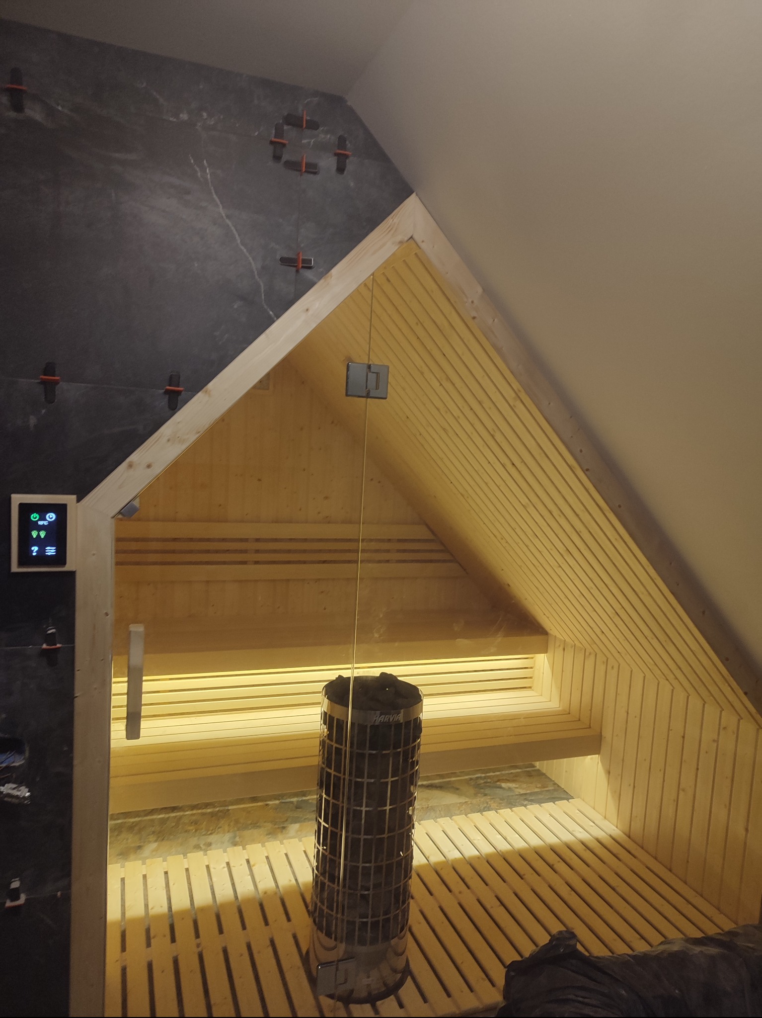 Wnętrze sauny z boazerią w kształcie trójkąta, piecem Harvia, czarną ścianą z płytkami i sterownikiem dotykowym, w trakcie montażu płytek widoczne krzyżyki dystansowe.