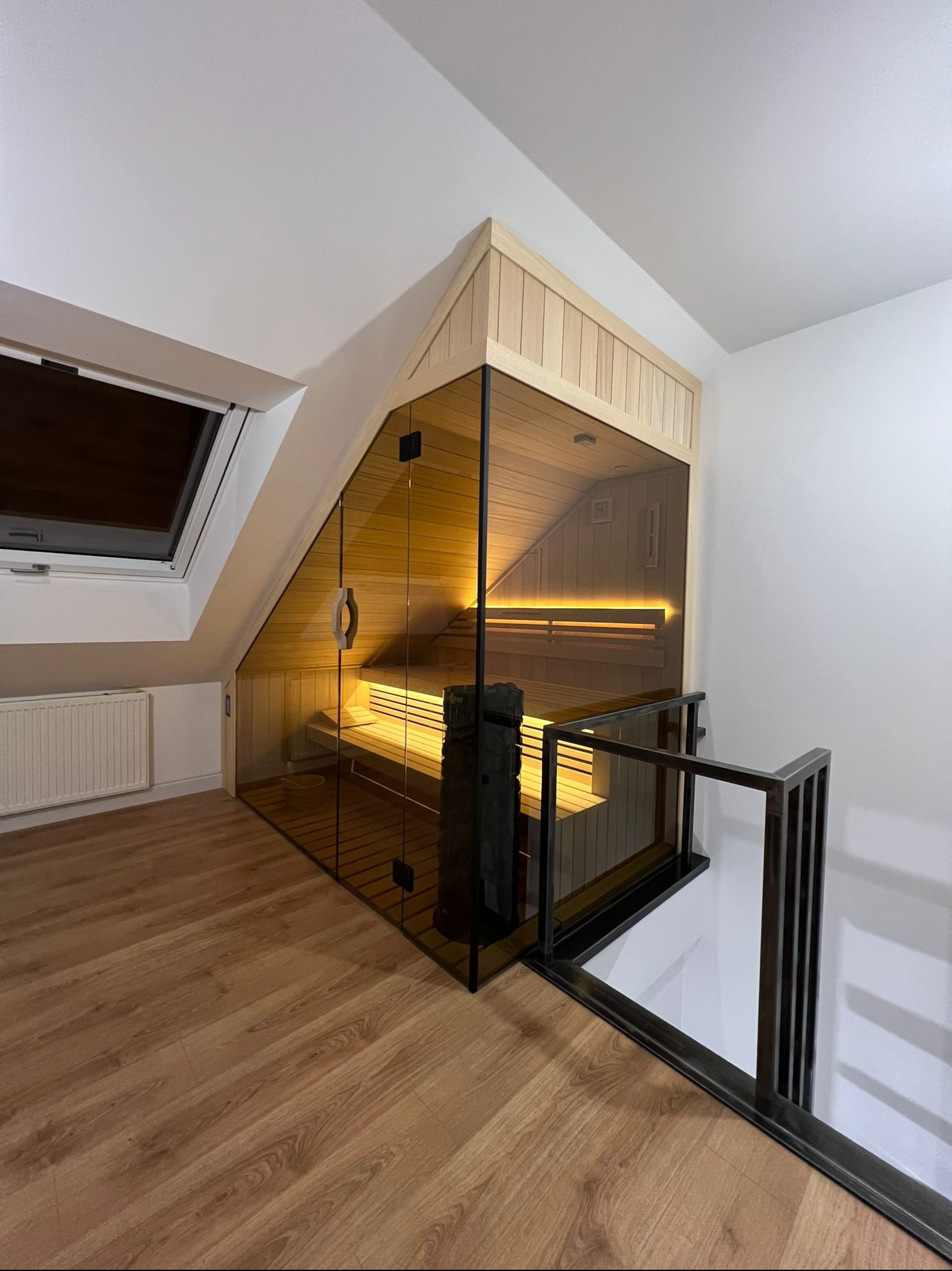 Sauna wkomponowana w poddasze, przeszklona ściana z drewnianym obramowaniem, widoczne ławy wewnątrz, oświetlenie LED, balustrada schodów z czarnego metalu.