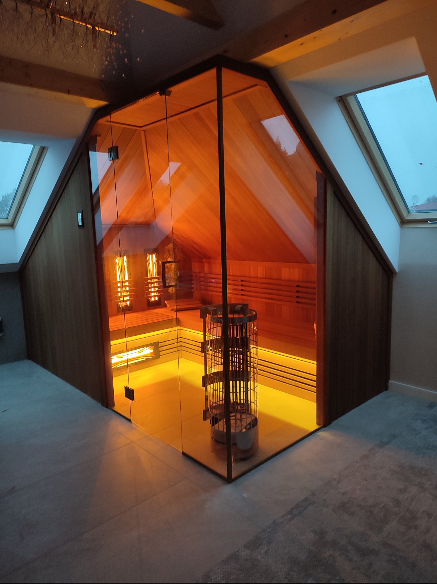 Nowoczesna sauna z przeszkloną ścianą, drewnianym wnętrzem i metalowym piecem w kształcie kosza, umieszczona na poddaszu z oknami dachowymi.