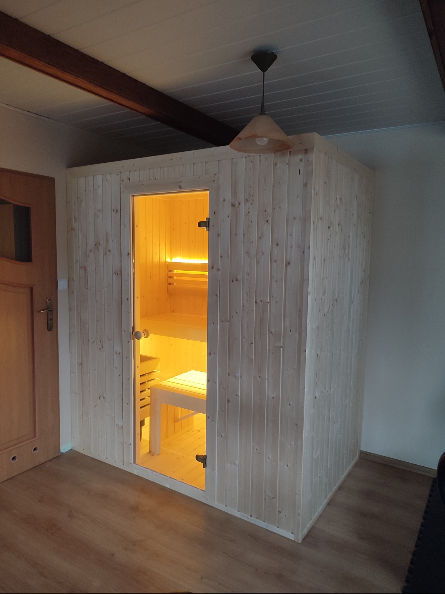 Wnętrze nowej, jasnej sauny z drewna, widoczne przez przeszklone drzwi, z ławkami i oświetleniem wewnątrz, umieszczonej w pomieszczeniu z drewnianą podłogą i lampą sufitową.