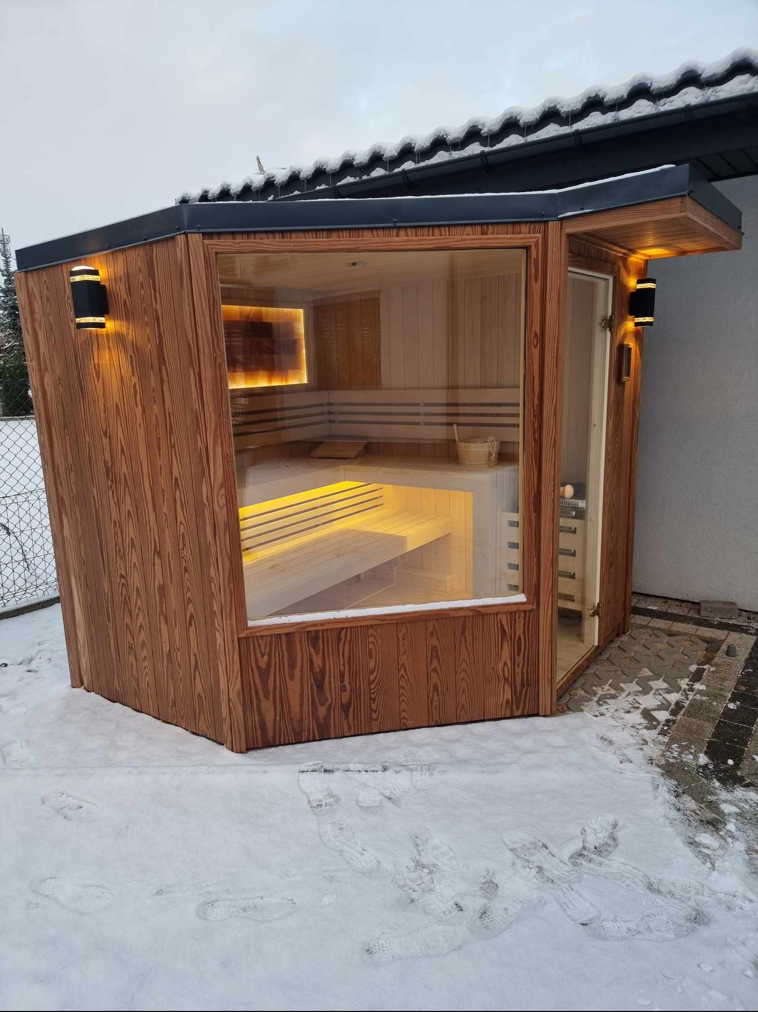 Drewniana sauna zewnętrzna z przeszkloną ścianą i oświetleniem, otoczona śniegiem, widoczna z zewnątrz.