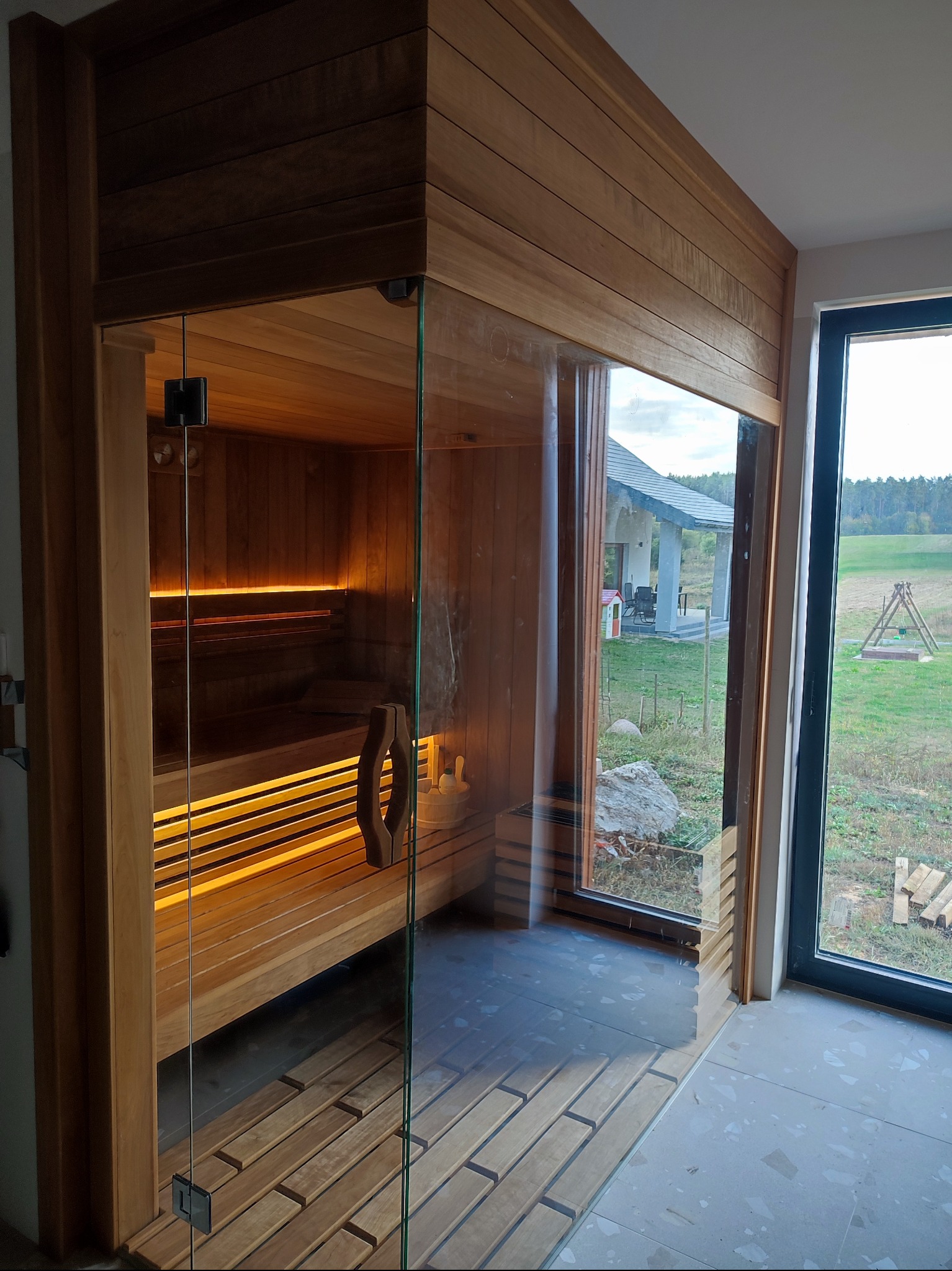 Nowoczesna sauna z przeszklonymi drzwiami, podświetlane drewniane ławki, widok na ogród z domem w tle, drewniane wykończenie ścian.