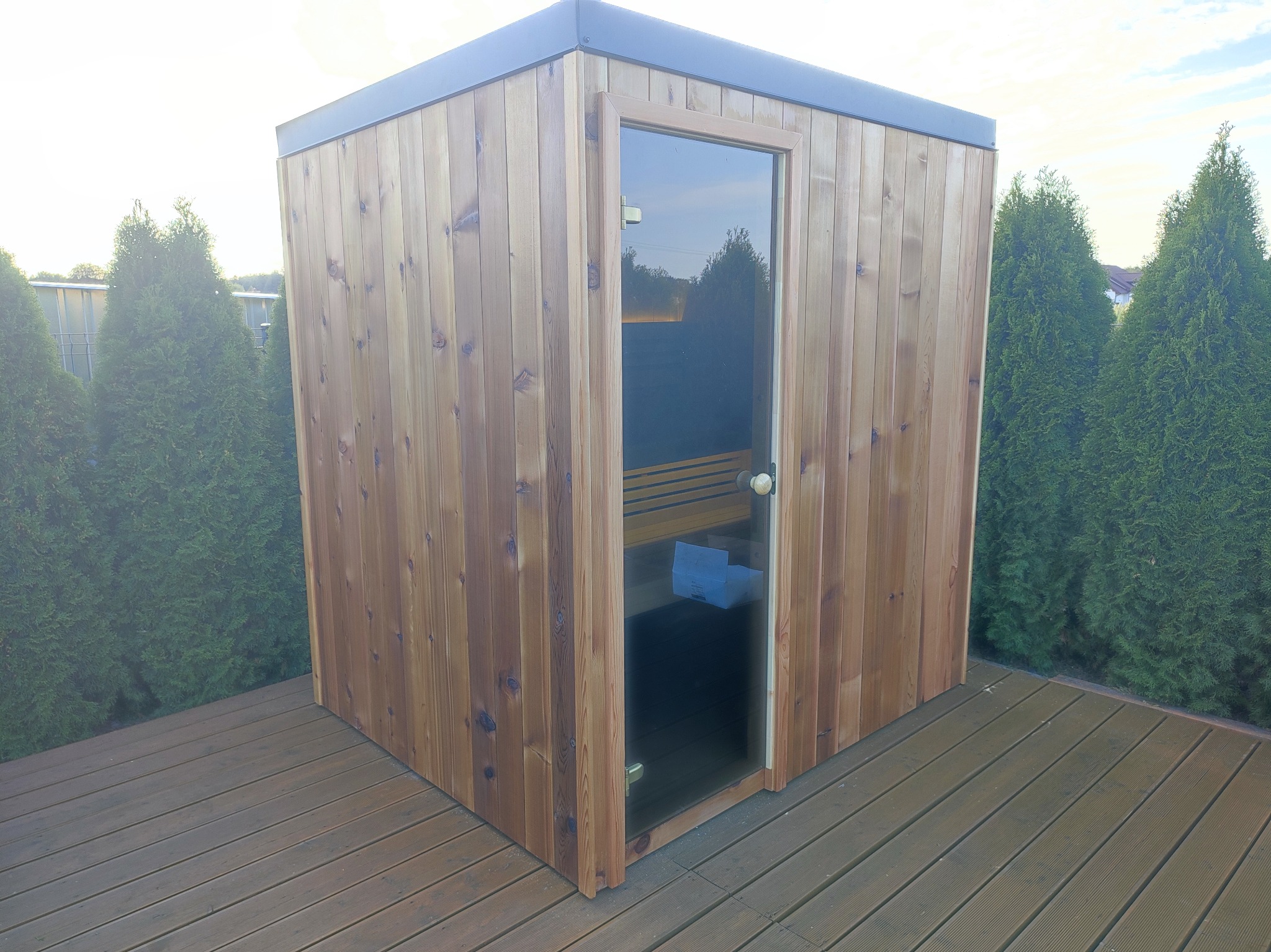 Drewniana sauna zewnętrzna z przeszklonymi drzwiami, umieszczona na drewnianym tarasie, otoczona tujami.