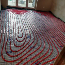 Czerwone rury ogrzewania podłogowego ułożone na folii z siatką, widoczne okno i fragment ściany, instalacja przed wylaniem posadzki.