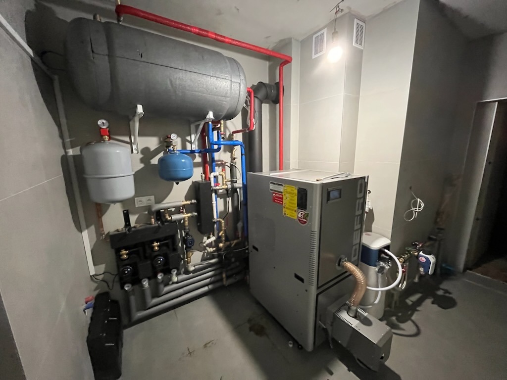 Zestaw urządzeń grzewczych w kotłowni: duży zbiornik akumulacyjny, piec z podajnikiem, naczynia wzbiorcze, rozdzielacz z pompami, liczne rury i zawory. Widoczne okablowanie elektryczne.