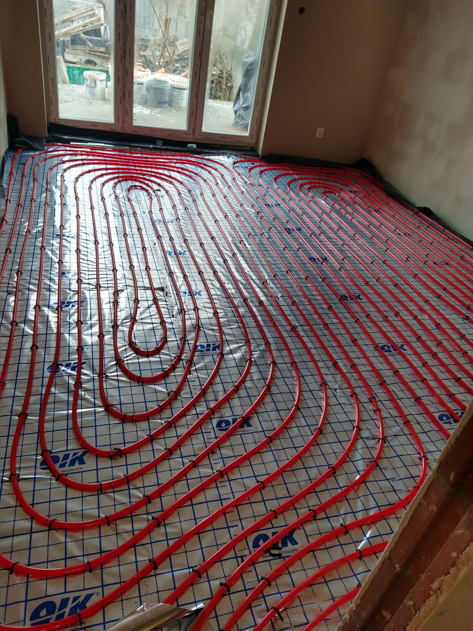 Czerwone rury ogrzewania podłogowego ułożone na folii z siatką, widoczne okno i fragment ściany, instalacja przed wylaniem posadzki.