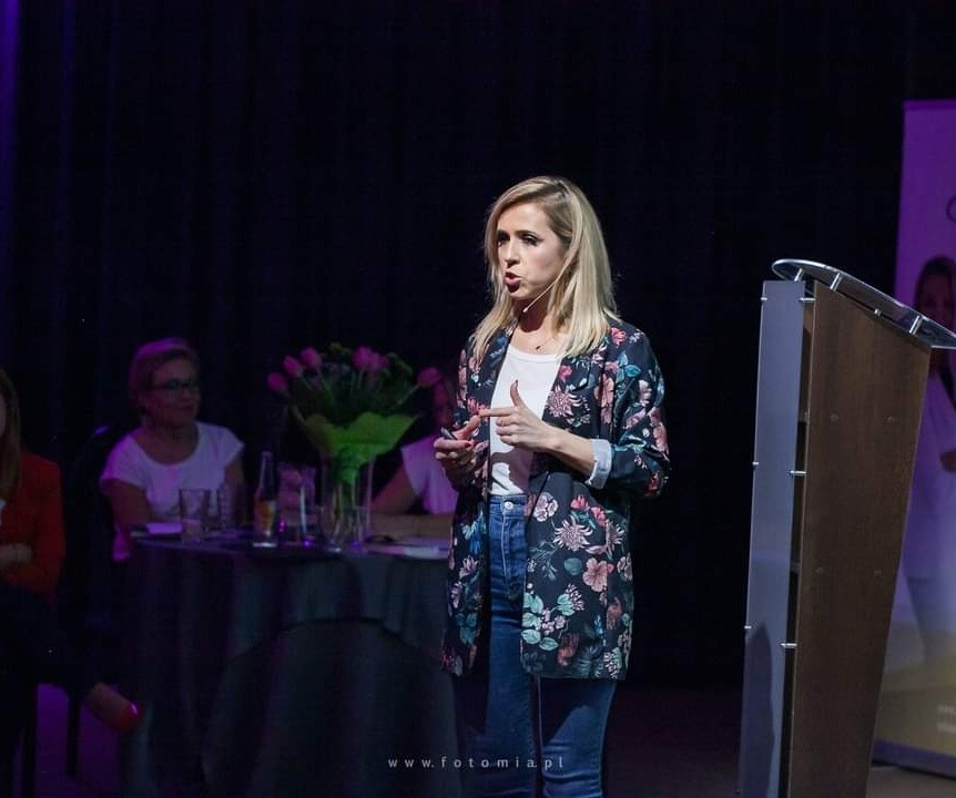 Kobieta w kwiecistej marynarce wygłasza prelekcję przy mównicy, w tle publiczność i bukiet tulipanów.