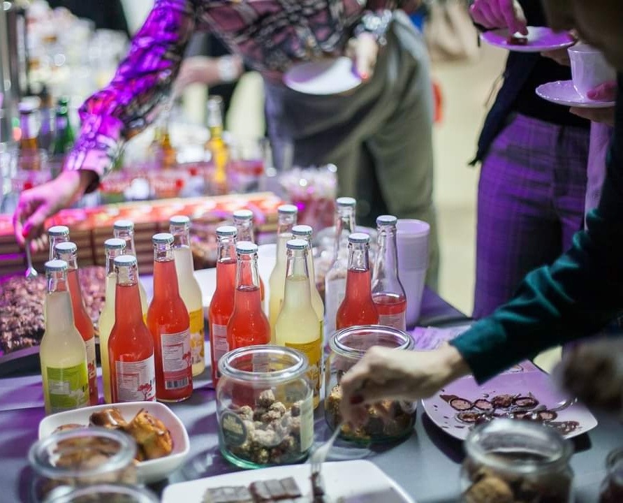 Bufet ze słodkimi przekąskami i napojami w szklanych butelkach, z widocznymi dłońmi osób sięgających po poczęstunek.