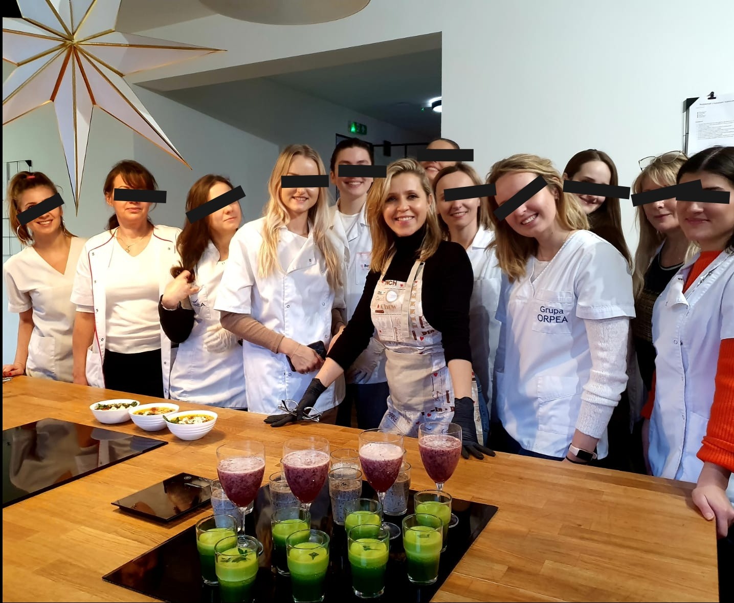 Grupa osób, w tym kobieta w fartuchu i czarnych rękawiczkach, prezentuje napoje i dania w miseczkach na drewnianym blacie. Część osób ubrana w białe uniformy z napisem 'Grupa ORPEA'.