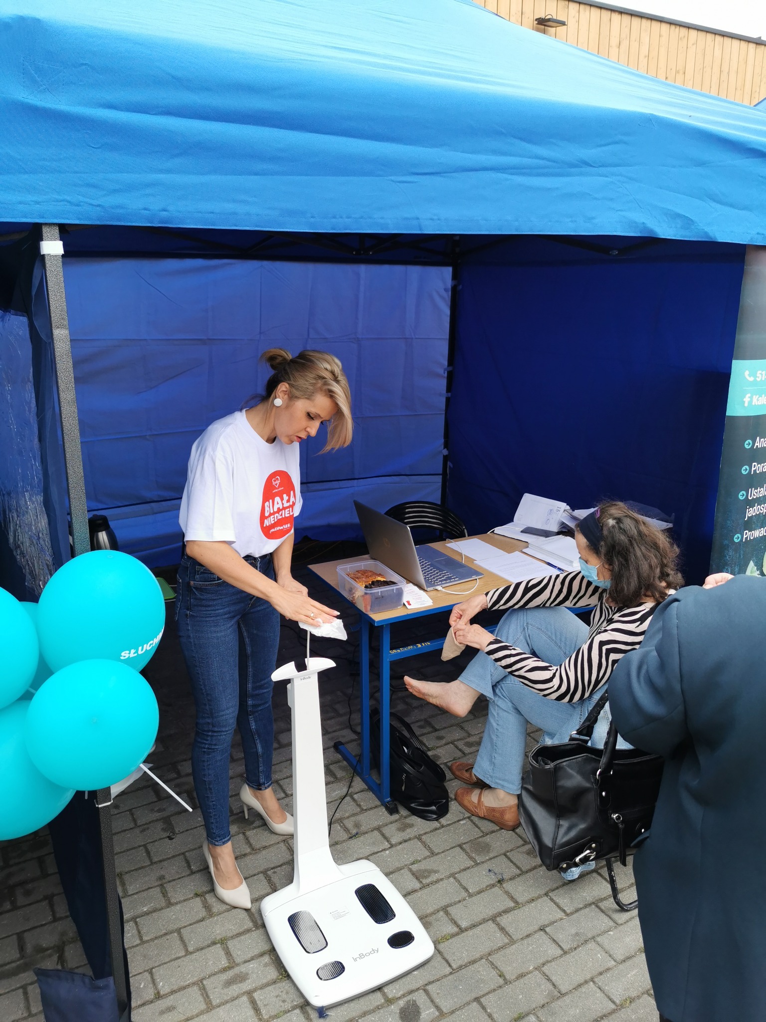 Kobieta w białej koszulce z logo czyści białą wagę InBody na tle niebieskiego namiotu, obok turkusowe balony i siedząca osoba w maseczce.