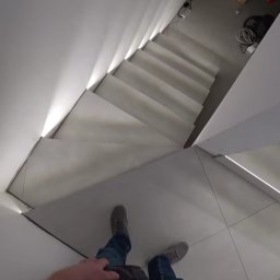 Kogucik - Wnętrze nowoczesnego domu z białymi schodami z podświetleniem LED, widok z góry na stopy osoby stojącej na kafelkach podłogowych.