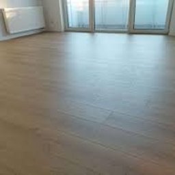 Kogucik - Jasne panele podłogowe w nowym, pustym pomieszczeniu z białymi ścianami i grzejnikiem, widoczne drzwi balkonowe z dostępem do światła słonecznego.