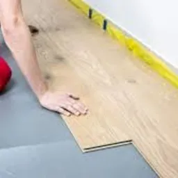 Montaż podłogi z paneli przez montera w spodniach roboczych z czerwonymi wstawkami, widoczne zabezpieczenie ściany taśmą malarską.