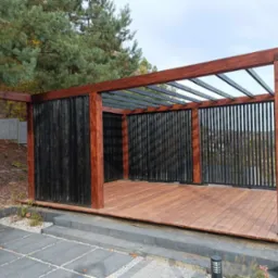 Nowoczesna altana z drewnianą podłogą i czarnymi pionowymi panelami, widok z zewnątrz na tle zieleni i kostki brukowej.