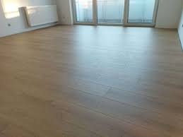 Jasne panele podłogowe w nowym, pustym pomieszczeniu z białymi ścianami i grzejnikiem, widoczne drzwi balkonowe z dostępem do światła słonecznego.