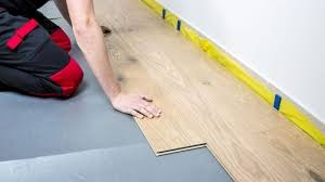 Montaż podłogi z paneli przez montera w spodniach roboczych z czerwonymi wstawkami, widoczne zabezpieczenie ściany taśmą malarską.