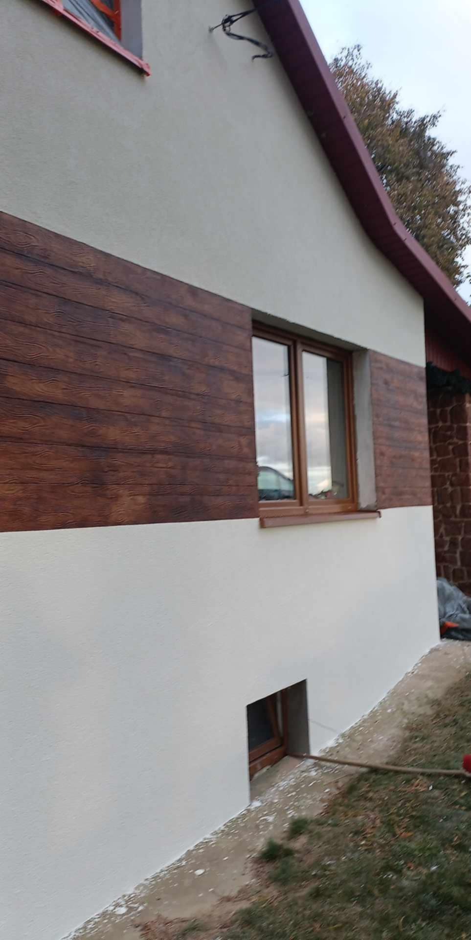 Elewacja domu z imitacją desek w kolorze brązowym na górnej części i białą farbą na dole, widoczne okno z brązową ramą i małe okienko piwniczne.