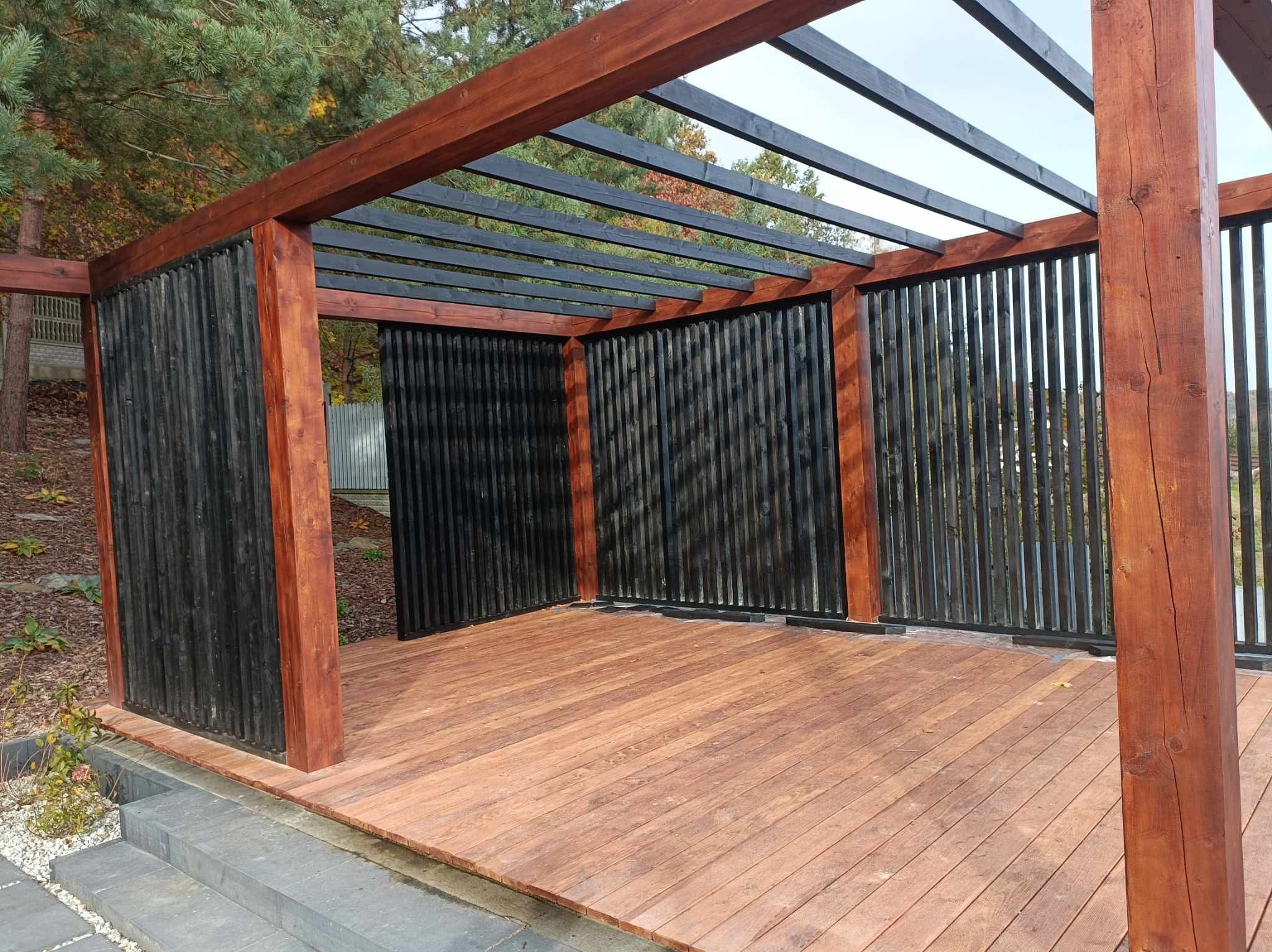 Drewniana konstrukcja z czarnymi panelami i brązowymi słupami, widok na taras z desek i schody z kostki brukowej, otoczona zielenią.