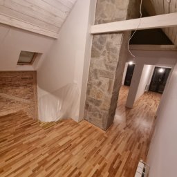 Grzegorz Wojdyła Gregor Bau - Wnętrze domu w trakcie remontu z drewnianą podłogą, kolumną obłożoną kamieniem, widoczną ścianą z cegły i zabezpieczoną folią, oraz fragmentem grzejnika.