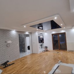 Grzegorz Wojdyła Gregor Bau - Wnętrze w trakcie prac wykończeniowych: ściana wyłożona cegłą dekoracyjną, drewniana podłoga, sufit z podwieszaną, lustrzaną częścią, widoczne narzędzia i osoba w tle.