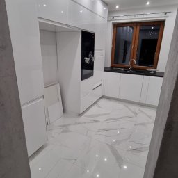 Grzegorz Wojdyła Gregor Bau - Nowoczesna, biała kuchnia z połyskiem w Łobozew Górny, z marmurową posadzką i oknem w drewnianej ramie. Widoczne oświetlenie sufitowe i wbudowany piekarnik.