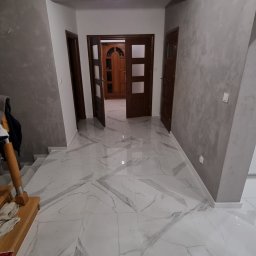 Grzegorz Wojdyła Gregor Bau - Wykończony korytarz z białymi marmurowymi płytkami podłogowymi, szarymi ścianami z efektem dekoracyjnym i ciemnymi drewnianymi drzwiami.