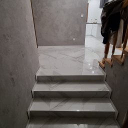 Grzegorz Wojdyła Gregor Bau - Wykończone schody z białego marmuru z szarymi żyłami prowadzące do przedpokoju z gładzią na ścianach i widokiem na białą kuchnię w tle.