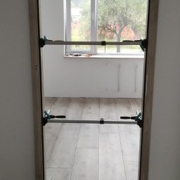 Grzegorz Wojdyła Gregor Bau - Ościeżnica drzwiowa mocowana rozpórkami teleskopowymi w nowym otworze, widoczny poziomica, karton i zabezpieczenie podłogi.