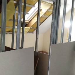 Grzegorz Wojdyła Gregor Bau - Stelaż metalowy pod płytę gipsowo-kartonową w poddaszu z oknem dachowym, widoczne płyty g-k przed montażem, żółta izolacja wewnątrz konstrukcji.