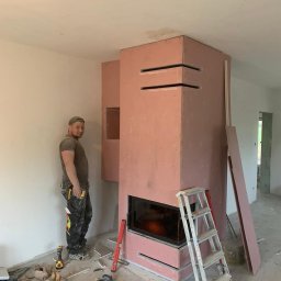 Grzegorz Wojdyła Gregor Bau - Monolityczna obudowa kominka z płyty karton gips w trakcie montażu, widoczny pracownik w ubraniu roboczym i drabina aluminiowa, na podłodze narzędzia i resztki materiałów.