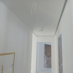 Grzegorz Wojdyła Gregor Bau - Korytarz w trakcie malowania na biało, widoczne kable wystające z sufitu, ściany przygotowane do malowania, zaklejone futryny drzwi.
